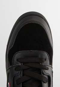 Baskets noires avec panneaux en cuir et en daim, lacets noirs et un petit drapeau rouge, blanc et bleu sur le côté.