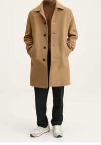 Beige Wollmantel mit Reverskragen, Frontknöpfen und Seitentaschen, kombiniert mit schwarzen Hosen und hellen Sneakers.