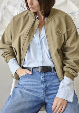 Donna che indossa una camicia azzurro chiaro abbottonata, una giacca bomber color kaki, una cintura nera e jeans blu, seduta con una mano in tasca.