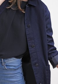 Chaqueta de botón en azul marino con un tejido texturizado, dos bolsillos frontales y un cuello clásico, superpuesta sobre una camiseta negra y unos jeans de mezclilla.