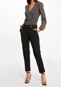 Blusa negra con lunares, mangas transparentes, escote en V profundo y cintura fruncida; combinada con pantalones negros a medida con detalles de botones dorados.