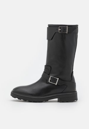 Botte en cuir noire mi-mollet avec deux boucles argentées, bout arrondi et semelle robuste, vue de côté gauche sur fond blanc.