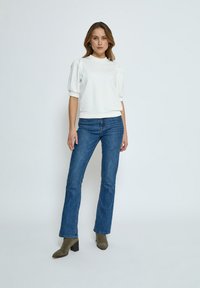 Vit topp med kort ärm och rund halsringning, med puffärmar. Kombinerad med blå utsvängda jeans och gröna klackade boots vid ankeln. Slät struktur överlag.