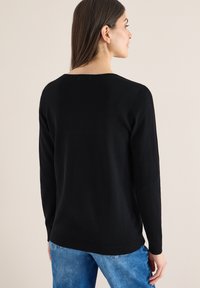 Femme aux longs cheveux noirs portant un pull noir à manches longues et un jean bleu, debout et tournée de dos sur un fond neutre.
