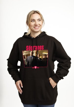 Smilende kvinde iført sort hættetrøje med "The Sopranos" logo og fire mænd i jakkesæt fra tv-showet på fronten. Hænderne i lommerne.