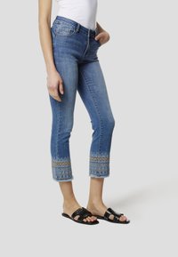 Lichtblauwe denim capri-jeans met een franjes zoom versierd met een patroon van borduurwerk in blauw en bruin. Gecombineerd met zwarte platte sandalen.