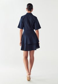 Femme avec un chignon portant des boucles d'oreilles en or, une robe bleu marine à volants et des sandales à talons beiges, se tenant devant un fond blanc.