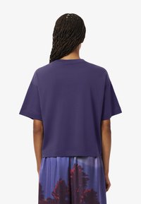 T-shirt di cotone viola con vestibilità rilassata, maniche corte e scollatura rotonda, mostrato da dietro con una texture liscia e morbida.