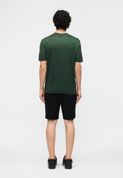 Άνδρας με σγουρά σκούρα μαλλιά, που φοράει σκούρο πράσινο πουκάμισο με κοντά μανίκια, μαύρο σορτς και μαύρα παπούτσια, στέκεται γυρισμένος προς τα πίσω σε λευκό φόντο.