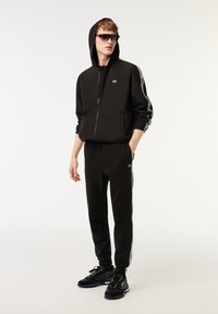 Svart idrottsdräkt med zip-up hoodie och joggebukser. Har vita sidostrick och grön logotyp. Svarta sneakers fulländar looken.