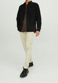 Camicia di velluto a costine nera con bottoni sopra un maglione marrone, abbinata a pantaloni color crema e scarpe nere stringate, in piedi contro uno sfondo chiaro.