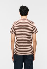 Άνδρας γυρισμένος με την πλάτη, φοράει ριγέ κοντομάνικη μπλούζα μπορντό και μπεζ και σκούρο μπλε τζιν, σε απλό λευκό φόντο.