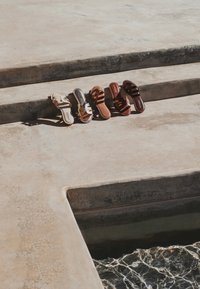 Cinq paires de sandales aux tons neutres disposées au soleil sur des marches en béton, à côté de l’eau ondulante.