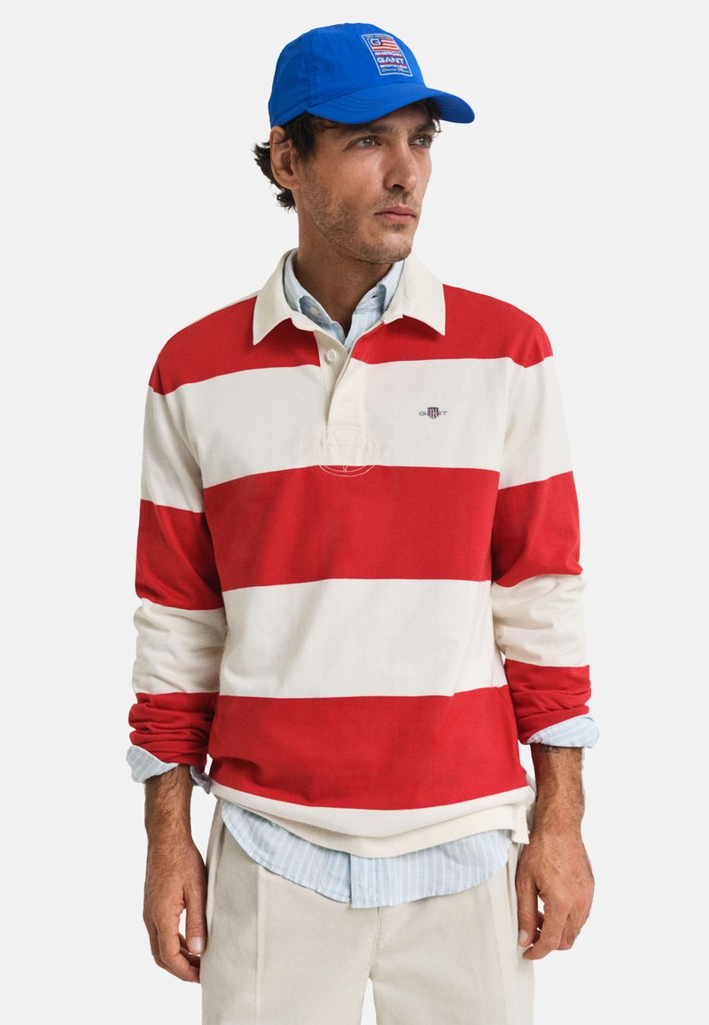 Rood-witte gestreepte polo met lange mouwen en een blauwe pet. Heeft een kraag en daaronder een lichtblauw geruit shirt in lagen.