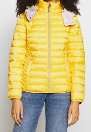 Person iført en skarpt gul quiltet jakke med lynlås foran og blå jeans mod en ensfarvet lys baggrund.