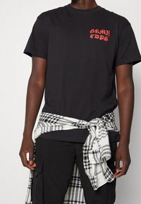 Černé bavlněné tričko s červeným grafickým textem na hrudi, doplněné bílou a černou kostkovanou flanelovou košilí uvázanou v pase.