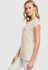 Beige Baumwoll-T-Shirt mit aufgerollten Kurzärmeln, das "Hoffnung" in bunten Buchstaben zeigt. Kombiniert mit weißen Hosen für einen lässigen Look.