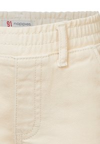 Pantalons en coton beige clair avec une finition texturée. Les caractéristiques incluent une taille élastique douce, des passants de ceinture et une petite étiquette de marque sur la taille.