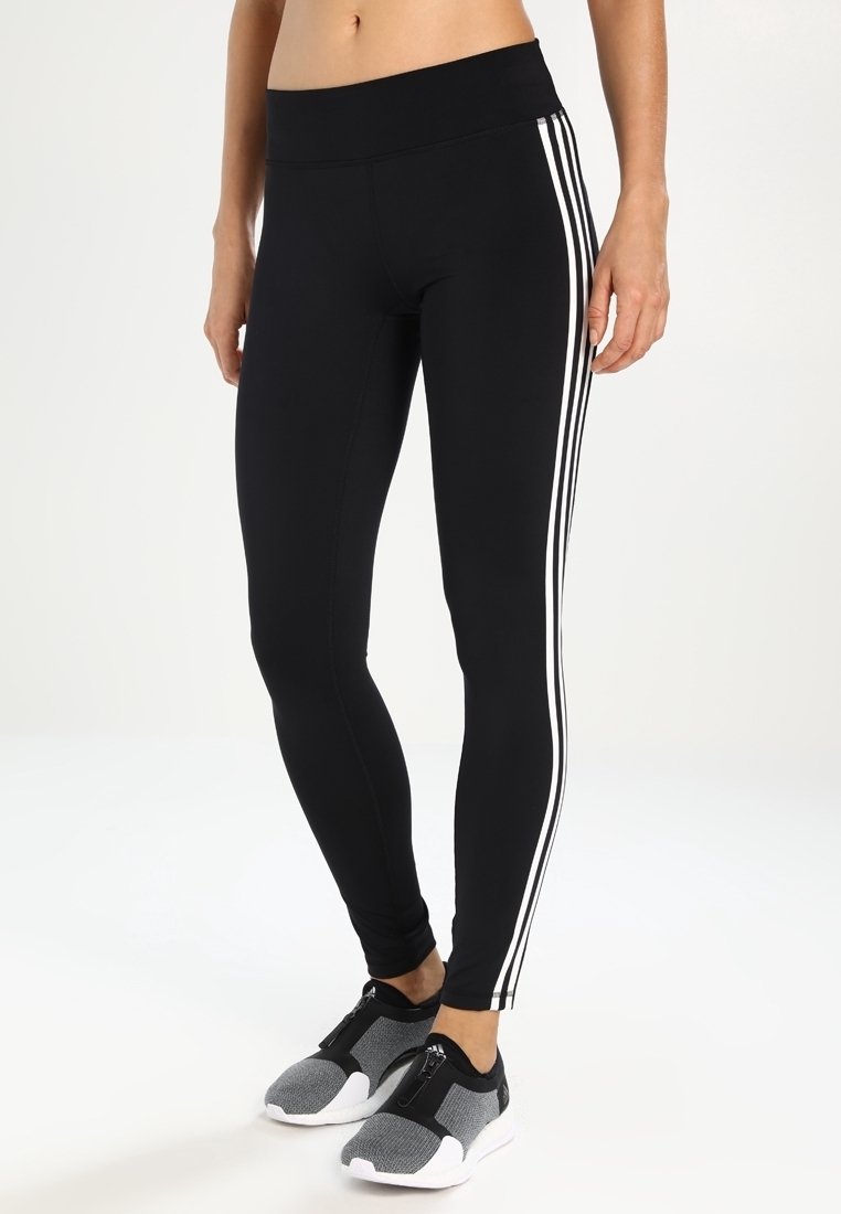Leggings negros con una cintura alta, que presentan dos franjas blancas a lo largo de los lados exteriores. Textura suave, ajuste ceñido. Combinados con zapatillas deportivas grises.