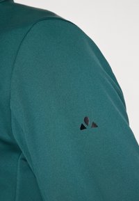 Grüne Stoffjacke mit glatter Textur. Besitzt ein schwarzes Logo auf dem linken Ärmel. Fokus auf das Ärmel-Design und die Gesamtform.