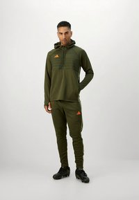 Olivzaļš džemperis ar kapuci un oranžām detaļām, kopā ar atbilstošām sporta biksēm. Abiem ir sportisks piegriezums un svītrains dizains augšdaļā.