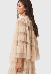 Beige tulle-klänning med lager av volanger och en ljus, luftig textur. Ärmar i elbow-längd och en lös, flödande siluett.