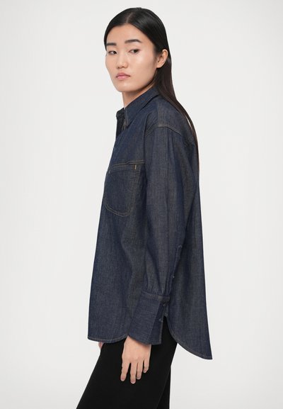 Tmavomodrá rifľová košeľa na zapínanie na gombíky, s náprsné vreckom, dlhými rukávmi a zakriveným lemom. Hladká textúra a minimalistický dizajn.