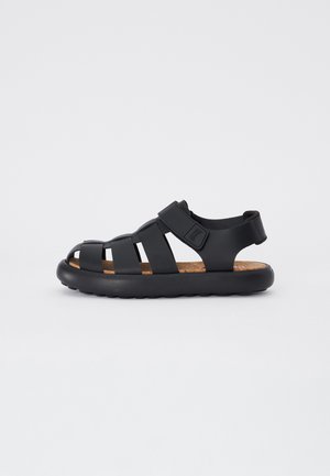 Black sandal made of rubber with open-toe design, featuring crisscross straps and a textured cork insole for comfort. Velcro strap for adjustment.