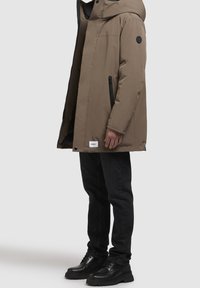 Chaqueta acolchada marrón con capucha, bolsillos laterales con cremallera y parches de logotipo en la manga; combinada con vaqueros negros y botas negras con cordones.