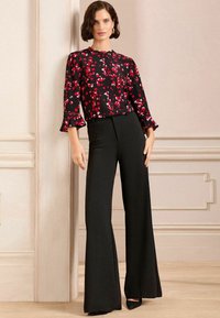 Schwarz-bluse mit floralen Mustern in Rot und Weiß, hat ausgestellte Ärmel; kombiniert mit hoch geschnittenen schwarzen Schlaghosen und spitzen schwarzen Absätzen.