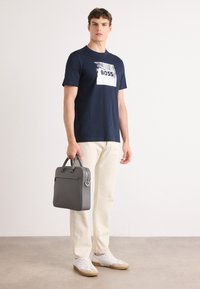 T-shirt blu navale con grafica bianca "BOSS", pantaloni crema, borsa strutturata grigia e sneakers bianche con suola color sabbia.