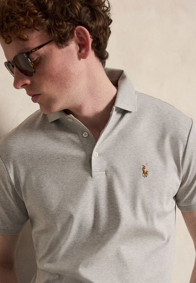Homme aux cheveux bouclés portant des lunettes de soleil foncées et un polo gris clair avec un petit logo brodé de joueur de polo sur la poitrine.