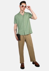 Camisa de botón verde de manga corta con un patrón texturizado, combinada con pantalones beige y zapatos marrones, realzada por unas grandes gafas de sol blancas.