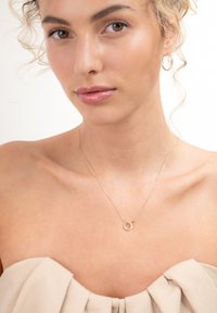 Gouden ketting met een ronde hanger, fijne ketting, gladde textuur en minimalistisch ontwerp. Model draagt een top in neutrale tinten.