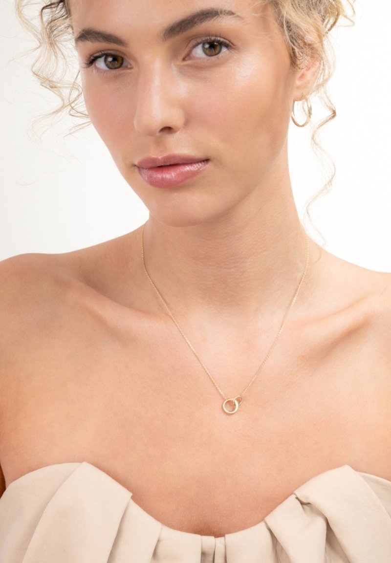 Gouden ketting met een ronde hanger, fijne ketting, gladde textuur en minimalistisch ontwerp. Model draagt een top in neutrale tinten.