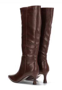 Botas marrones hasta la rodilla, hechas de cuero suave, con puntera afilada y tacón cónico delgado, presentando una textura suave y arrugada en el eje.