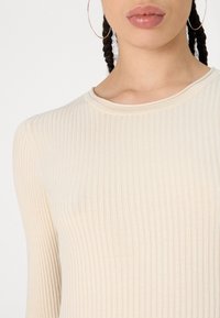 Haut beige à manches longues côtelé avec un col rond et un tissu texturé, présentant de fines lignes verticales et une coupe ajustée.