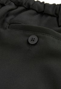 Tissu noir avec un bouton proéminent à quatre trous, à côté d'une couture de poche. Le matériau semble texturé et durable.