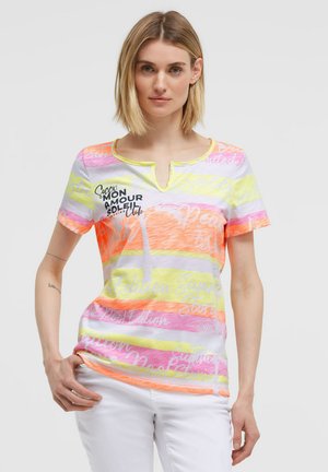 Woman with shoulder-length blonde hair wearing a short-sleeve colorful striped T-shirt and white pants, standing with one hand in pocket.