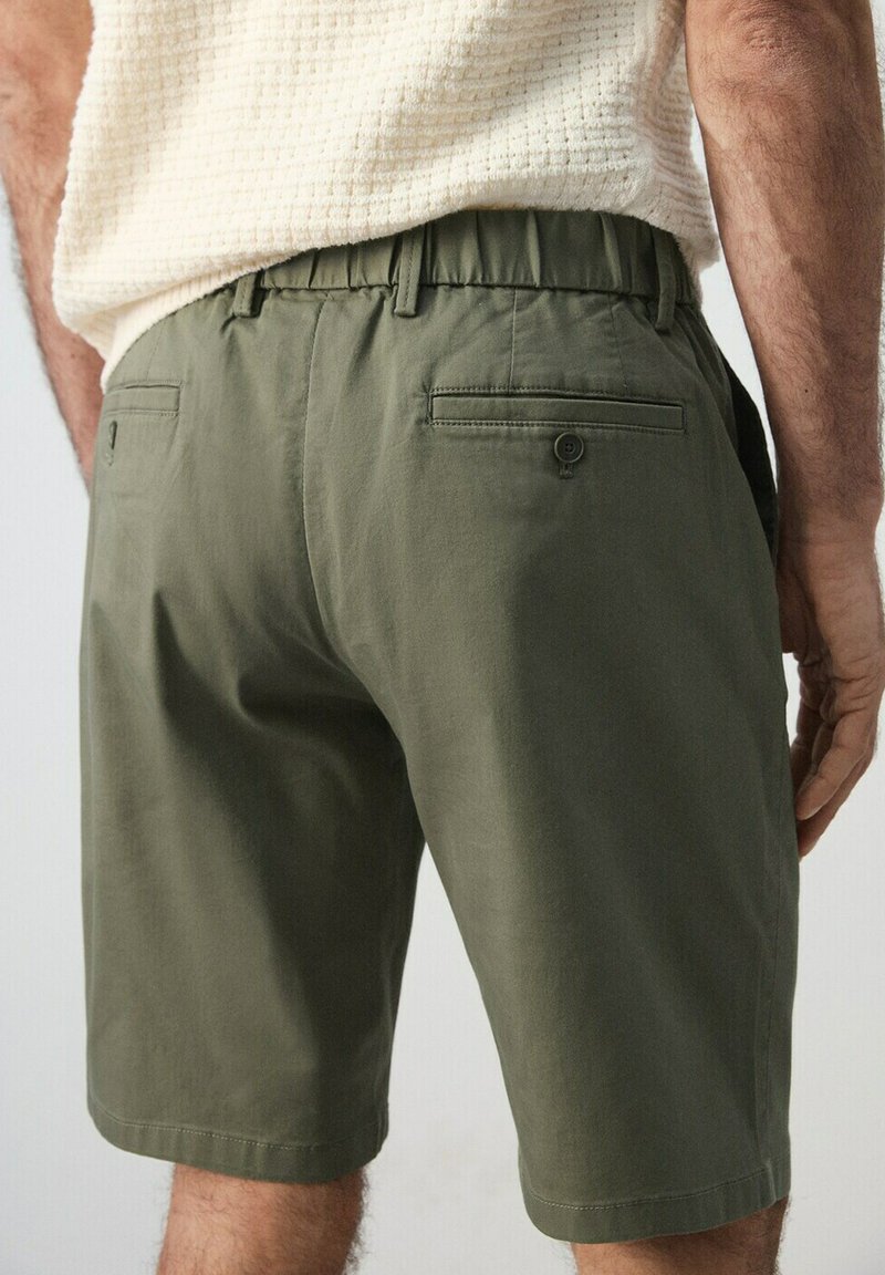 Homme portant un short vert olive avec des poches arrière boutonnées et une taille élastique, assorti à une chemise crème texturée.