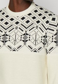 Gebreide trui met een crèmekleurige basis en zwarte geometrische patronen, een ronde halslijn en geribbelde manchetten voor een textuurafwerking.