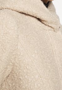 Beige fleece frakke med en tekstureret overflade, der har en bred krave og en let oversized pasform, som fremviser syninger ved siden af.
