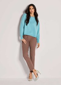 Frau mit langen dunklen Haaren, die einen hellblauen Pullover, braune Hosen und mehrfarbige High Heels trägt, steht an einer hellen Wand.