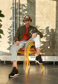 Jeune homme en veste verte, chemise orange, jean et baskets assis sur des chaises colorées empilées, à l'intérieur avec de la lumière du soleil et des ombres de plantes sur le mur.