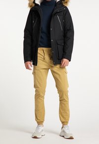 Mężczyzna w czarnej kurtce parka z futrzanym kapturem, granatowym golfem, beżowych spodniach typu jogger cargo i białych trampkach stoi na białym tle.