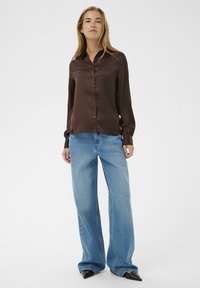 Cămașă din satin maro cu butoni, cu mâneci lungi, asortată cu blugi evazați de un albastru deschis și pantofi cu toc ascuțit negri. Design simplu și curat.