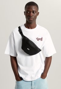 Zwarte heuptas met afgeronde vorm, verstelbare riem en logopatch, gedragen over een wit T-shirt. Lichtblauwe spijkershorts zichtbaar.