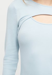 Gaiši zils garu piedurkņu tops ar horizontālu izgriezumu tieši zem atslēgas kaula, atsedzot neskartu ādu.