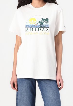 T-shirt blanc Adidas avec un logo représentant une scène de plage avec des palmiers, l'océan, le soleil et un voilier, porté avec un jean bleu.
