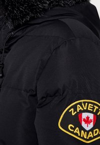 Chaqueta acolchada negra con un cuello de piel sintética, que presenta un parche redondo con "ZAVETTI" y una hoja de arce roja en la manga.